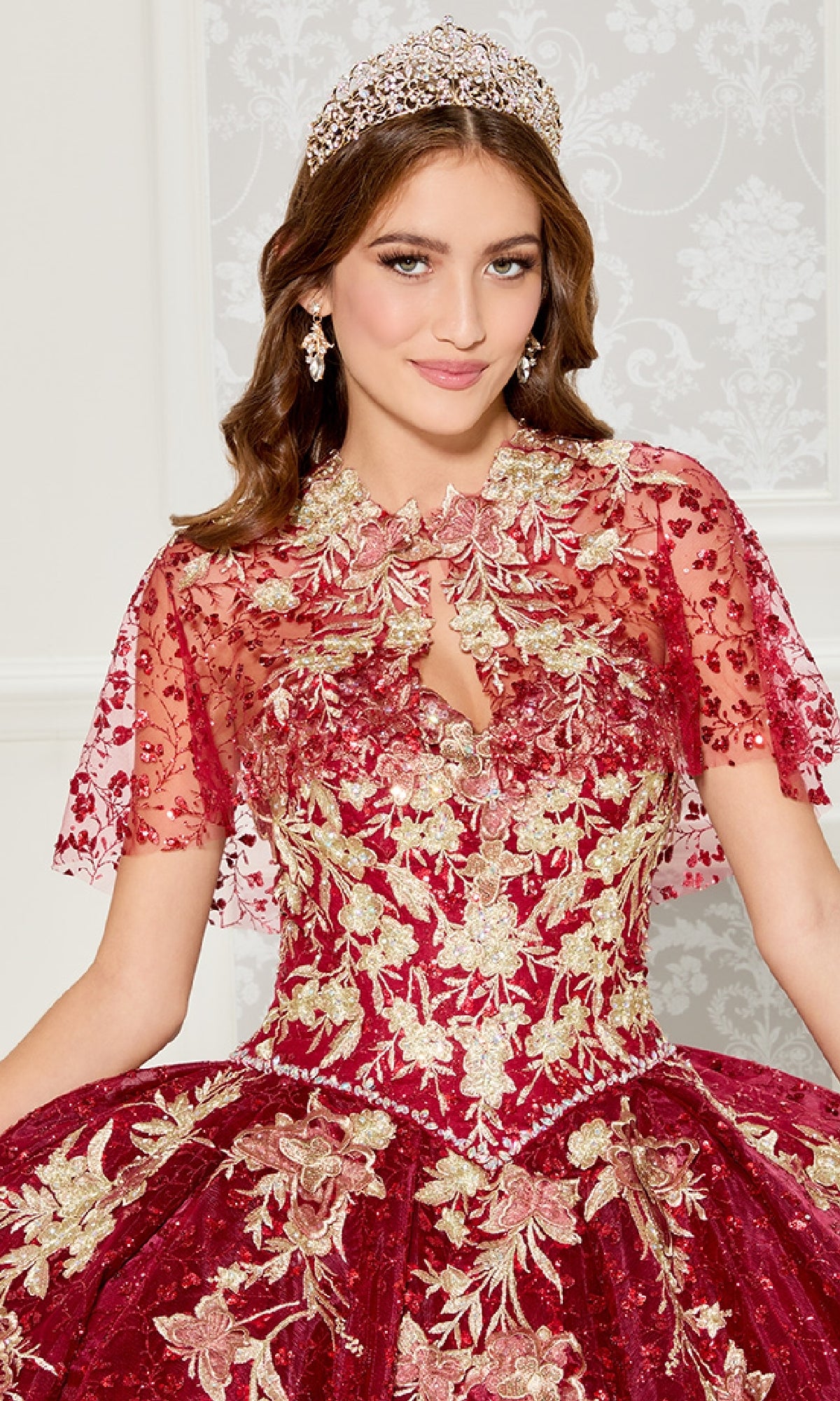 A woman poses indoors against a light patterned wall, wearing the Princesa PR30121 Strapless Quinceanera Dress in red and gold embroidery, accessorized with a tiara and earrings.